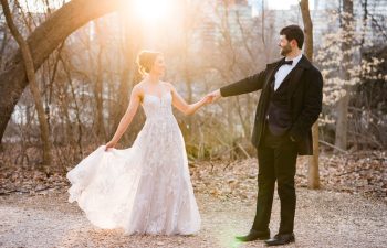 A bride in a white wedding dress and a groom in a dark suit hold hands and smile at each other outdoors in a wooded area with sunlight shining behind them.