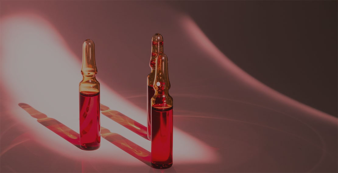 Two glass ampoules filled with red liquid standing upright on a smooth surface with pink and red light reflections.