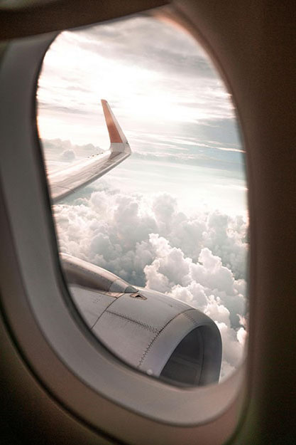 View from an airplane window showing the wing, engine, and clouds below with sunlight breaking through the sky.