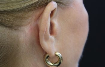 Close-up of a person’s left ear wearing a thick, gold hoop earring; the person has light skin and blonde hair pulled back.