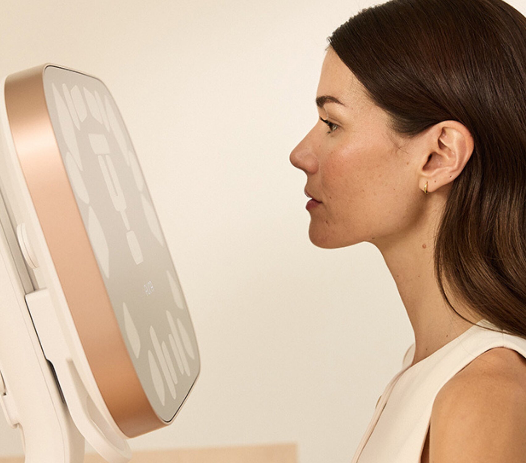 Side-profile view of a person standing close to a large, square body-composition scanning device mounted on a stand. The person faces the screen with a neutral expression in a softly lit indoor setting. The device has a light-colored frame with a rose-gold edge and abstract markings on its display.