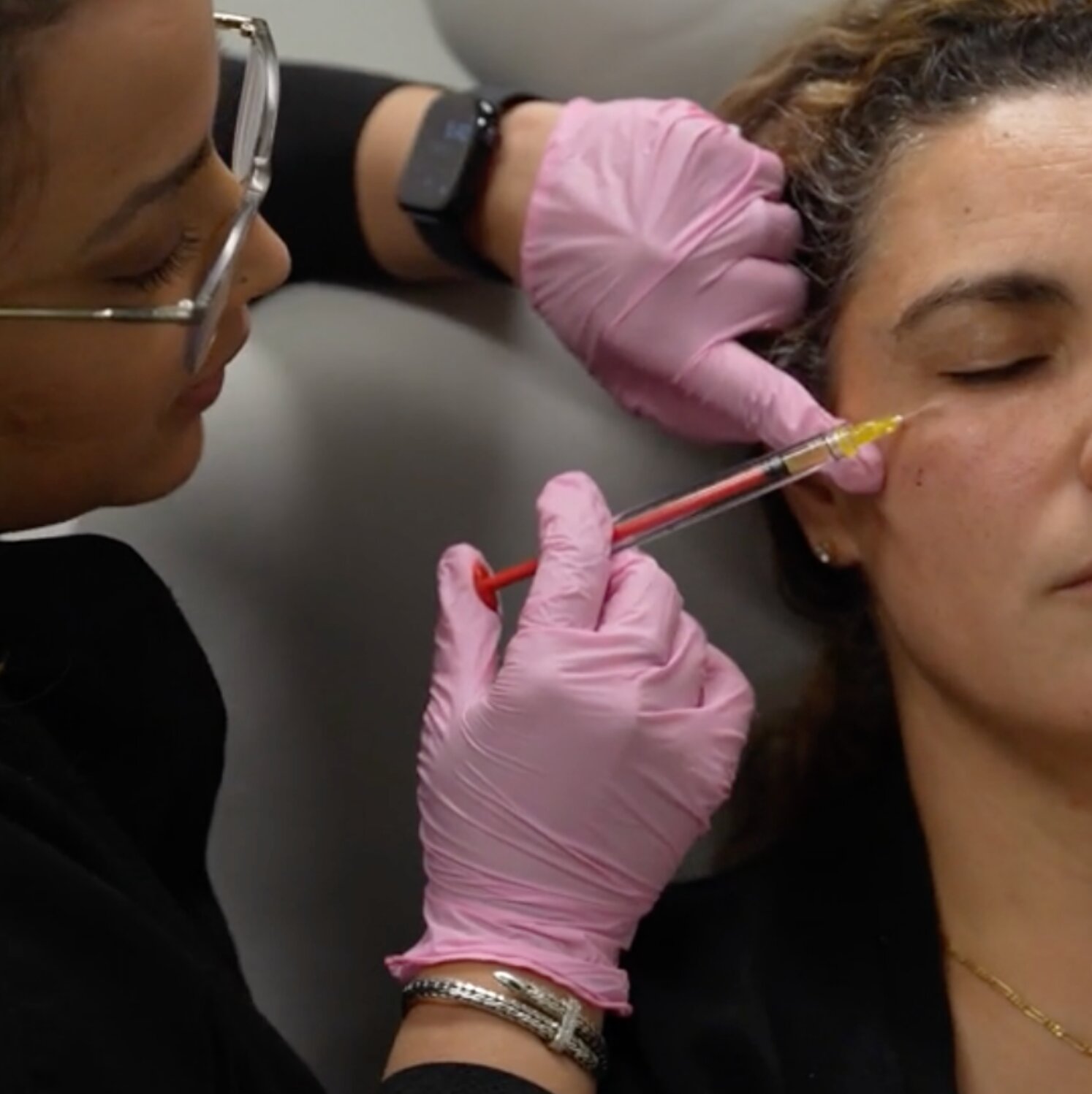 A medical or aesthetic professional wearing pink nitrile gloves and clear-framed glasses administers a facial injection using a cannula near a seated patient's cheekbone area. The patient has her eyes closed. The practitioner is also wearing a smartwatch and bracelet.